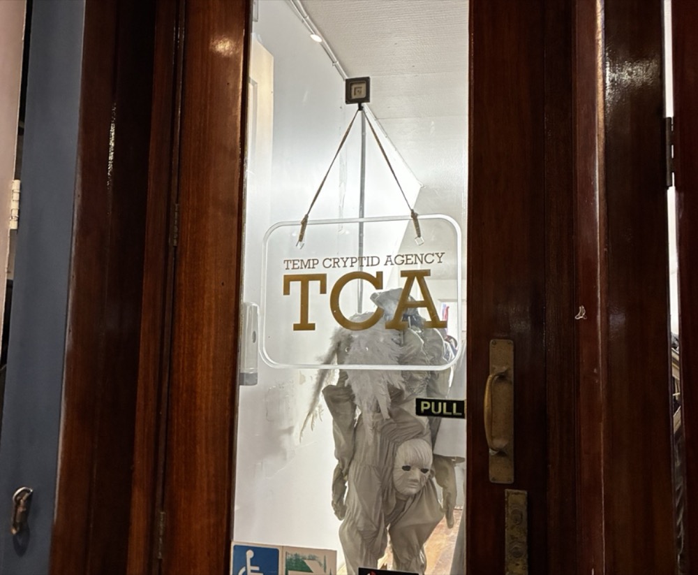 A wooden door with a hanging clear sign reading "TEMP CRYPTID AGENCY / TCA" in gold, a pale masked costume figure behind the glass and a PULL sticker by the handle